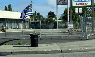 A picture of a black barrel with boards on the top, a flag pole, and a thin blue line flag attached. The barrel has several thick black wires connected to it, one runs along the sidewalk