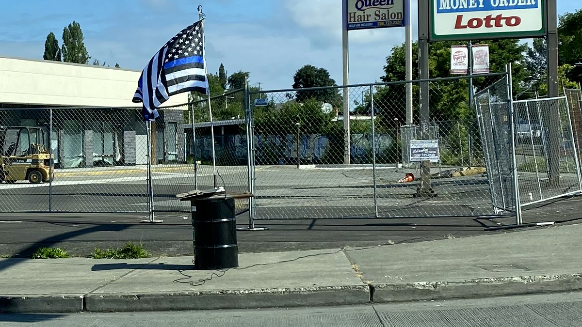 A picture of a black barrel with boards on the top, a flag pole, and a thin blue line flag attached. The barrel has several thick black wires connected to it, one runs along the sidewalk