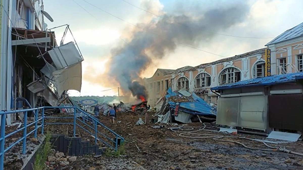 Downtown Bakhmut, Ukraine after a Russian artillery barrage is in ruins with buildings on the main street heavily damaged. Debris fills the road and in the background, a fire burns with a column of smoke rising in the air. A small group of civilians are in the center of the picture, walking away from the camera through the debris in the street