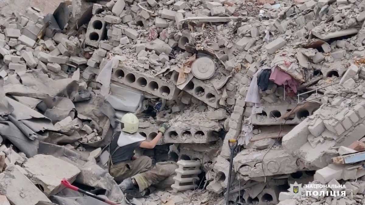 A searcher is in a massive pile of rubble in Chasiv Yar, looking between sections of concrete, rebar and bricks - clothing and other belongings are scattered in the debris