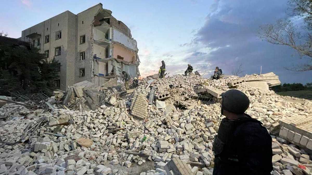 A firefighter looks at a partially collapsed block of apartments have a Russian missile attack in Chasiv Yar. The light brown stone and brick apartment block possibly six stories high before the attack, is collapsed. Three searchers are in the distance, standing on a pile of rubble 10 to 15 meters high, half of the building remains standing in the background.