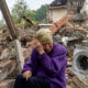 An 83 old woman dressed in purple with a her wrapped sits in the foreground weeping. Behind her is the destroyed home of her neighbors who were killed in a Russian missile attack. Among the debris is the belongings of her neighbors, including a shattered washing machine, clothing, and household goods