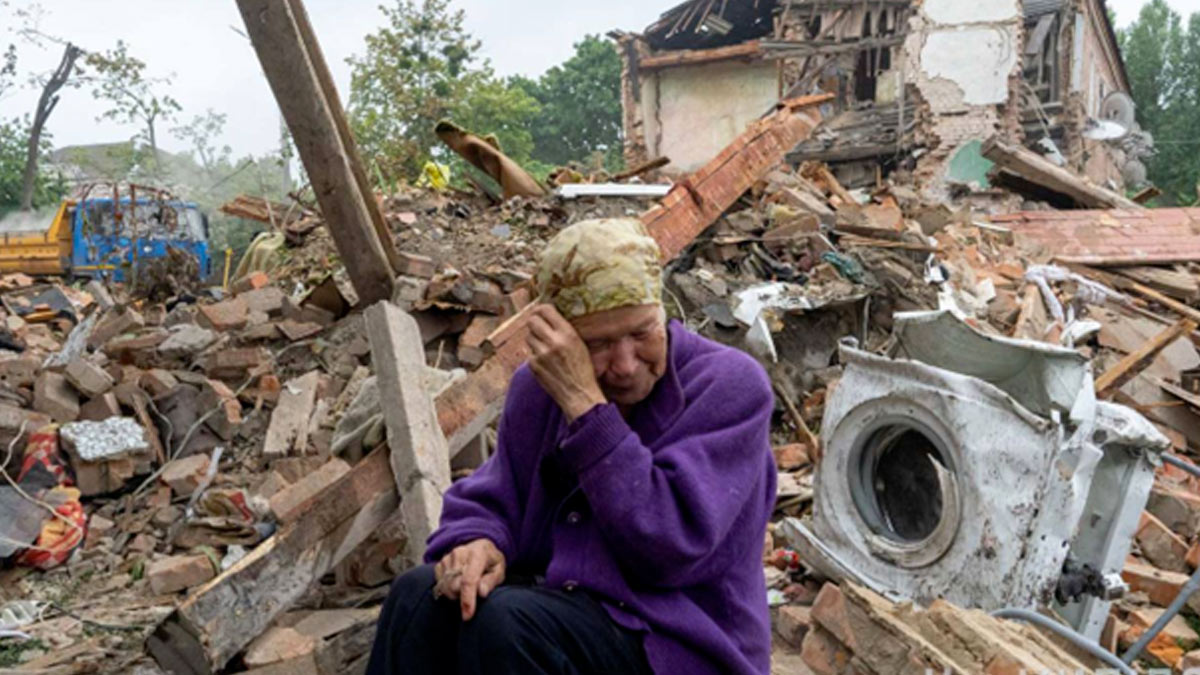 An 83 old woman dressed in purple with a her wrapped sits in the foreground weeping. Behind her is the destroyed home of her neighbors who were killed in a Russian missile attack. Among the debris is the belongings of her neighbors, including a shattered washing machine, clothing, and household goods
