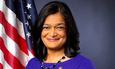 United States Congressperson Pramila Jayapal wearing a blue dress, and a necklace with black stones, an American flag is over her right shoulder