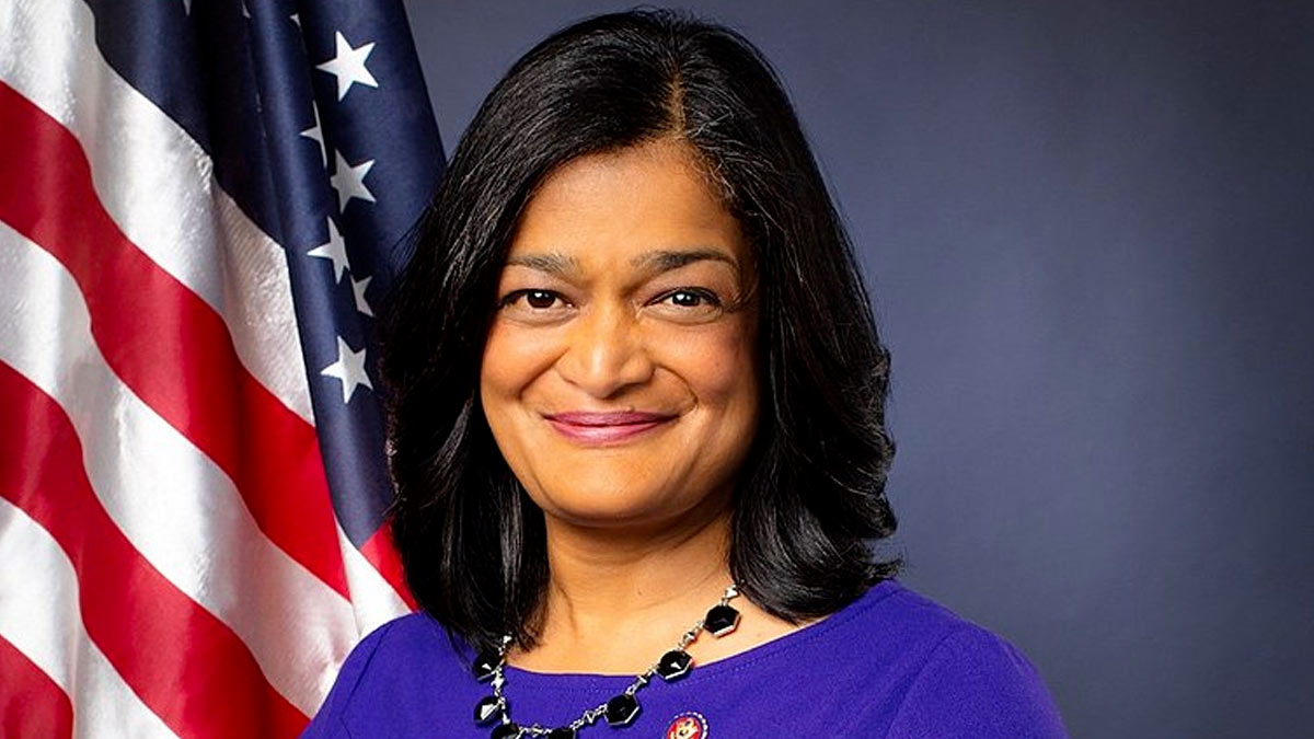 United States Congressperson Pramila Jayapal wearing a blue dress, and a necklace with black stones, an American flag is over her right shoulder