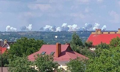 Some rises on the horizon as the 1st Army Corps shells Ukrainian positions in Avdiivka. The red roofs of Donetsk are in the foreground with the town of Avdiivka out of sight over a ridge, it is midday in summer with hazy clouds in the sky and lush trees