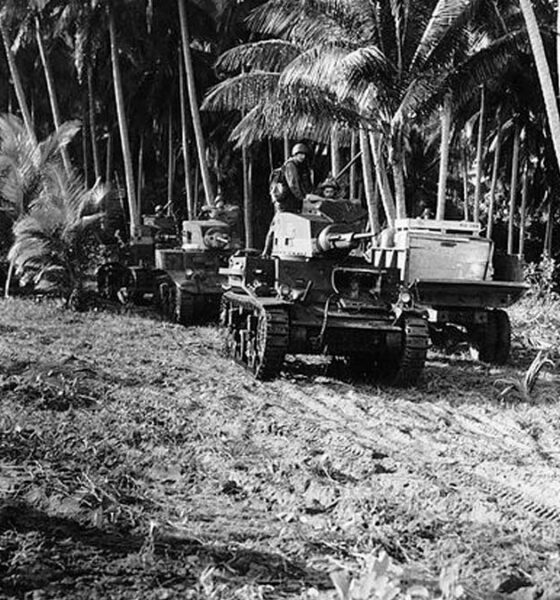 A black and white photo from 1942 taken on Guadalcanal during World War II shows American M2 light tanks and a supply truck on a makeshift road by a grove of trees.