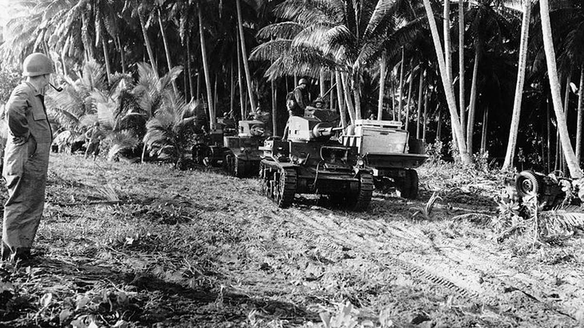 A black and white photo from 1942 taken on Guadalcanal during World War II shows American M2 light tanks and a supply truck on a makeshift road by a grove of trees.