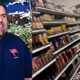 A split screen photo of Jeff Leiwis, a former Juanita High School security guard. On the left is Lewis in 2012 when he worked for the school, he is wearing a dark blue shirt and holding a walkie talkie smiling. On the right is Lewis in 2022 in Target in the condiment aisle, pushing a shopping cart wearing a dark blue t-shirt