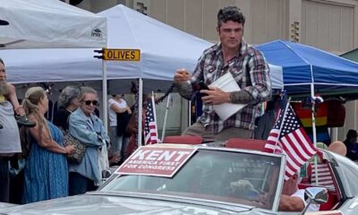 Joe Kent, a GOP Congressional primary candidate for the Washington 3rd District sits on the roof of a C3 Corvette Stingray in silver, wearing a flannel style shirt and looking off at the crowd. A sign that says in white letters with a red background Kent America First for Congress is taped on the windshield, people are looking in on from pop up tents along the side of the road