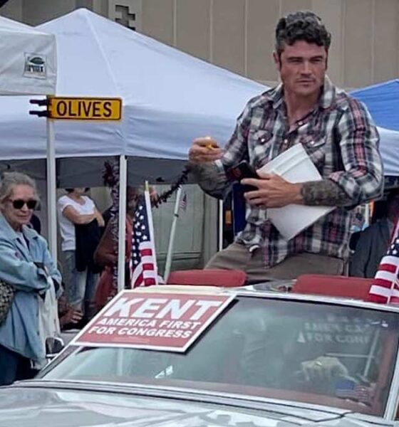 Joe Kent, a GOP Congressional primary candidate for the Washington 3rd District sits on the roof of a C3 Corvette Stingray in silver, wearing a flannel style shirt and looking off at the crowd. A sign that says in white letters with a red background Kent America First for Congress is taped on the windshield, people are looking in on from pop up tents along the side of the road