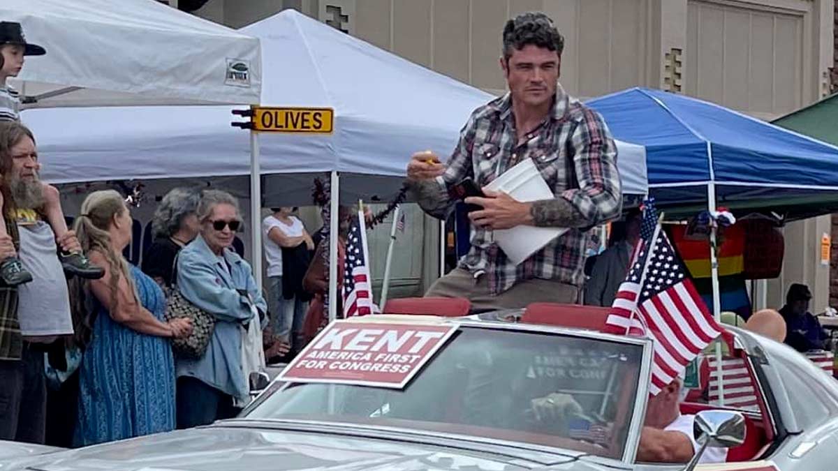 Joe Kent, a GOP Congressional primary candidate for the Washington 3rd District sits on the roof of a C3 Corvette Stingray in silver, wearing a flannel style shirt and looking off at the crowd. A sign that says in white letters with a red background Kent America First for Congress is taped on the windshield, people are looking in on from pop up tents along the side of the road