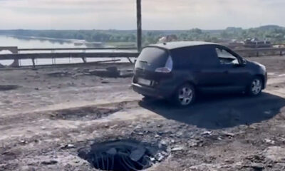 A compact car drives across a bridge that has been moderately damaged in a rocket attack. There are craters all through the bridge deck, with enough undamaged space for a single lane of traffic. Debris strews the surface with Dnipro River in the background. The car is driving very slowly.