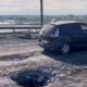 A compact car drives across a bridge that has been moderately damaged in a rocket attack. There are craters all through the bridge deck, with enough undamaged space for a single lane of traffic. Debris strews the surface with Dnipro River in the background. The car is driving very slowly.