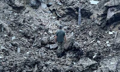 A man stands in a crater that is about 5 meters deep after a Russian cruise missile destroyed a water tower in western Ukraine. The ground is gray-black from the attack