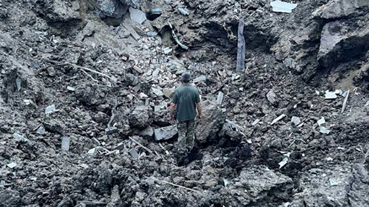 A man stands in a crater that is about 5 meters deep after a Russian cruise missile destroyed a water tower in western Ukraine. The ground is gray-black from the attack