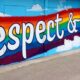 A mural on a wall in Olympia with the words Respect and Love in white text with a black outline on a rainbow colored background. The mural is surrounded in sky blue with puffy clouds above and below