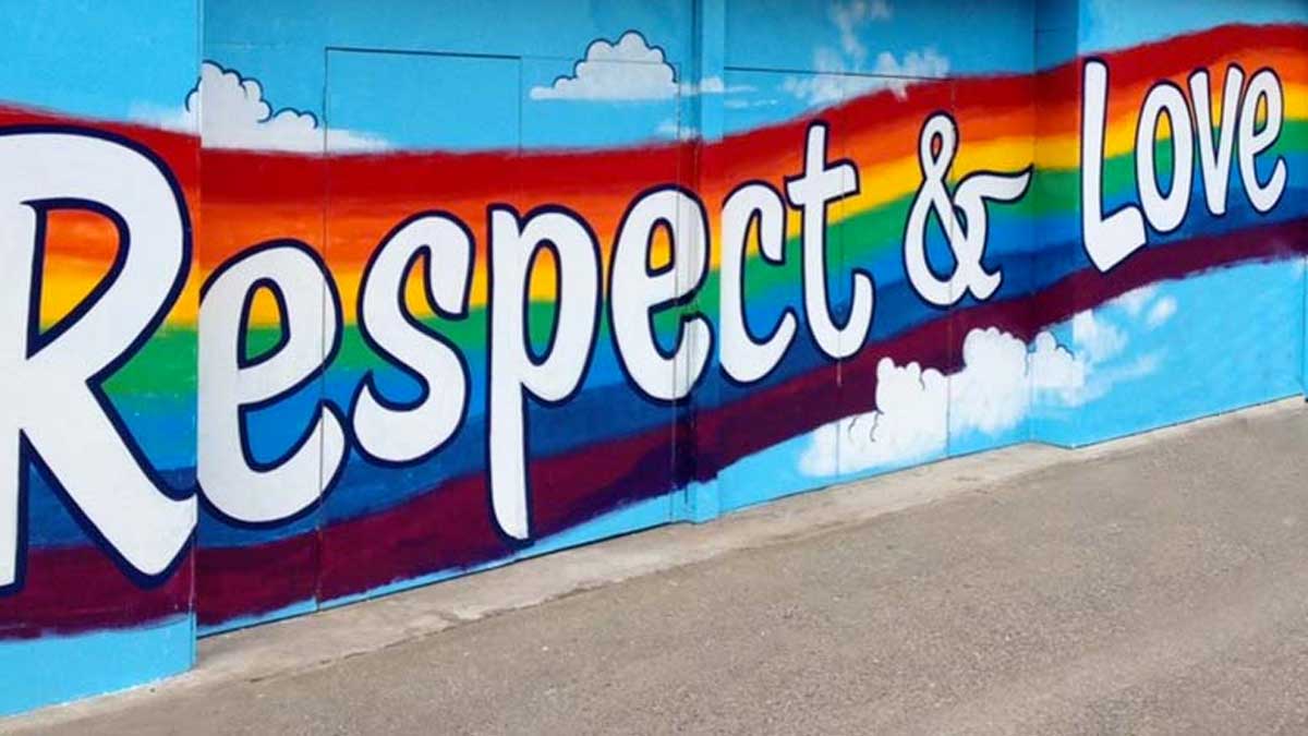A mural on a wall in Olympia with the words Respect and Love in white text with a black outline on a rainbow colored background. The mural is surrounded in sky blue with puffy clouds above and below