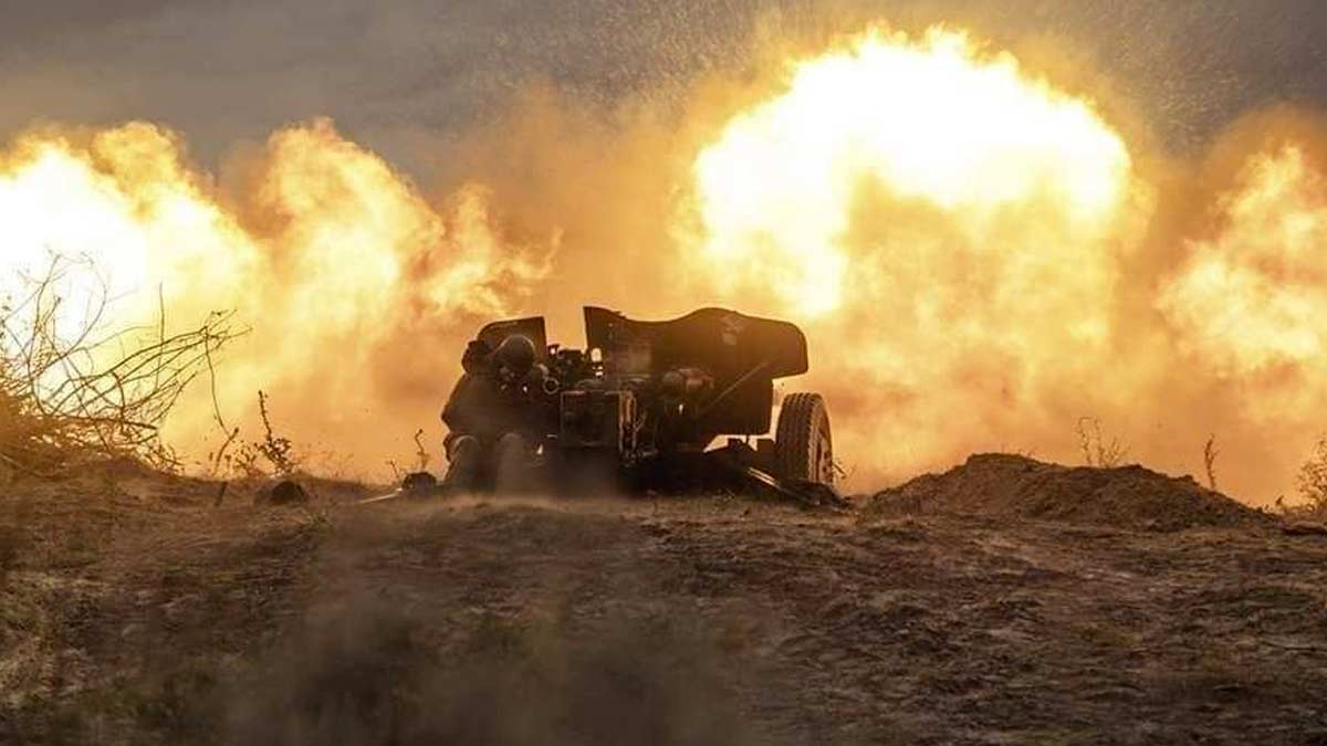 A soldier fires a Soviet-era D30 artillery piece in an unknown location in the Luhansk oblast of Ukraine. The ground has been pulverized by battle with nothing green or living in the foreground or background. The photo captures the moment the artillery piece fires, with the soldier hunched over, ears covered. There is an orange fireball that has illuminated the smoke orange. The artillery piece may be used for direct fire, with a low angle and what appears to be an explosion close to the position