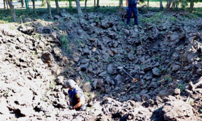 A large crater caused by a missile attack in Kharkiv that is around 6 meters deep with dark chunks of earth over turned. A police man stands at the bottom and a shovel lays on the ground