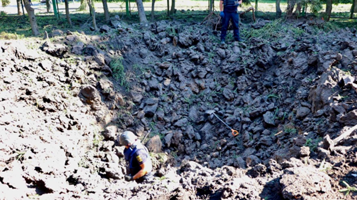A large crater caused by a missile attack in Kharkiv that is around 6 meters deep with dark chunks of earth over turned. A police man stands at the bottom and a shovel lays on the ground