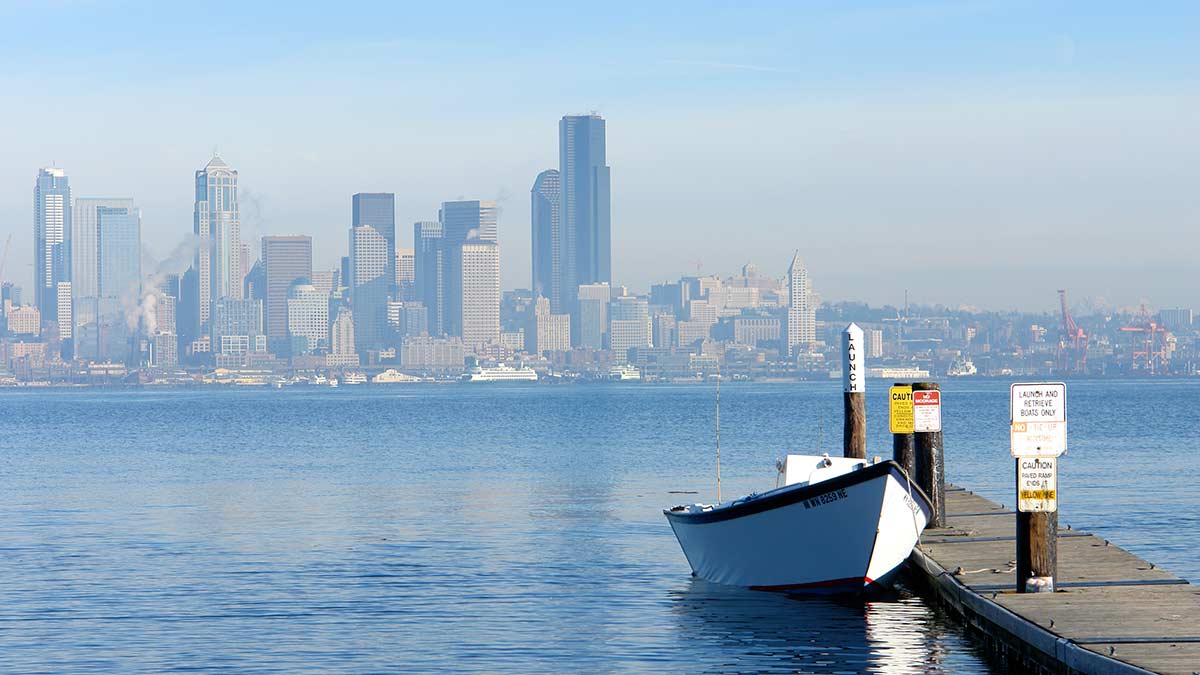 The Seattle skyline as viewed from across Elliott Bay is slightly obscured in summer haze. A rowboat is tied to a narrow floating dock in the foreground. The Columbia tower is at the center of the image, with the Port of Seattle just visible on the right