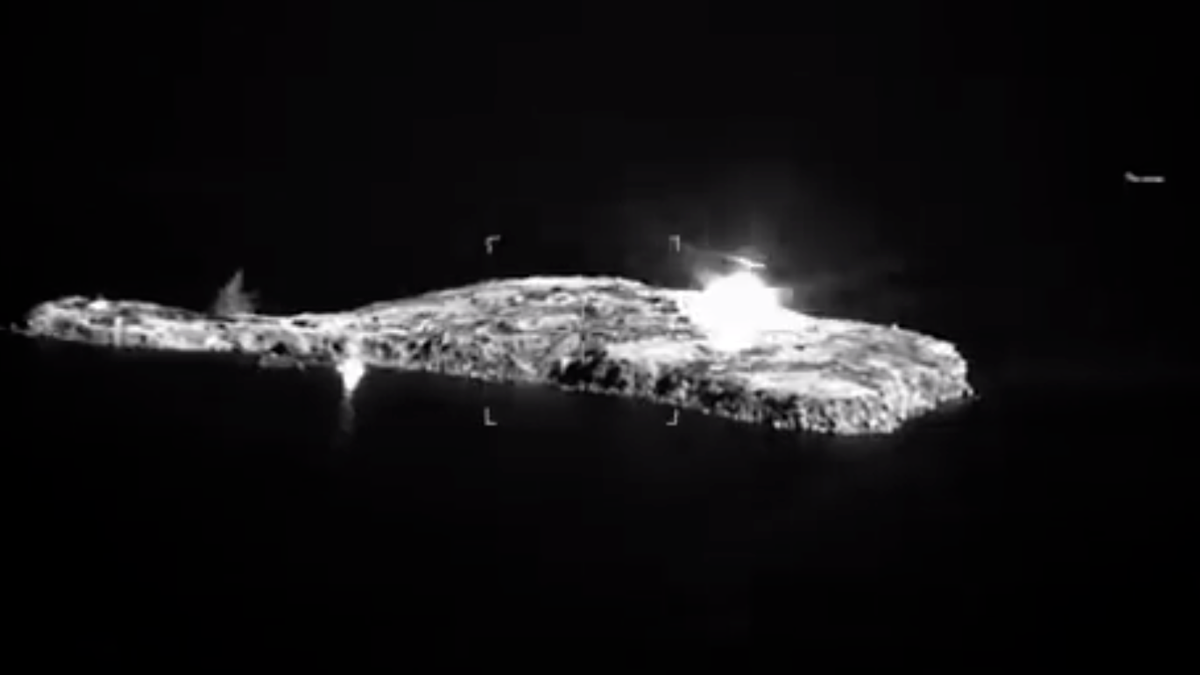 A black and white still image from a drone video, showing snake island in the black sea being bombed by a russian su-30 aircraft. There are two bombs that landed in the ocean with splashes in the image and one bomb exploding on the island