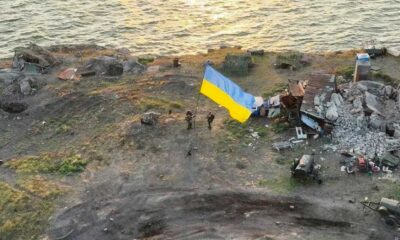 A drone takes a picture of several Ukrainian soldiers on Snake Island at sunrise. The ocean can be seen near by. A very large Ukrainian flag has been raised with destroyed Russian military equipment in the background.