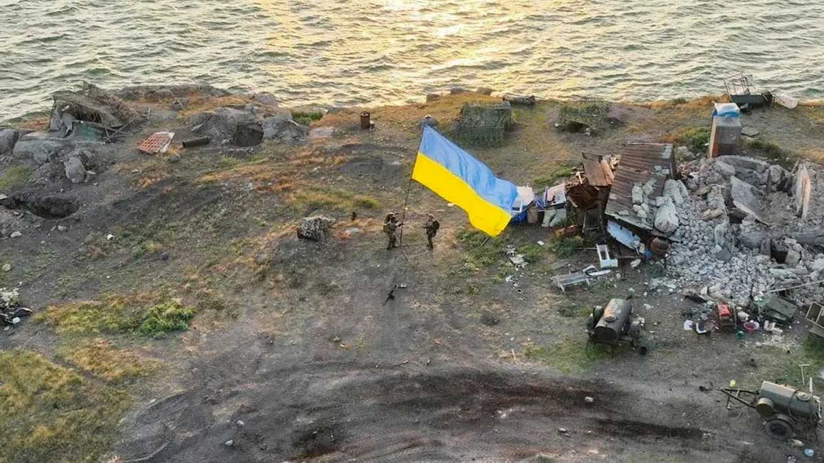 A drone takes a picture of several Ukrainian soldiers on Snake Island at sunrise. The ocean can be seen near by. A very large Ukrainian flag has been raised with destroyed Russian military equipment in the background.