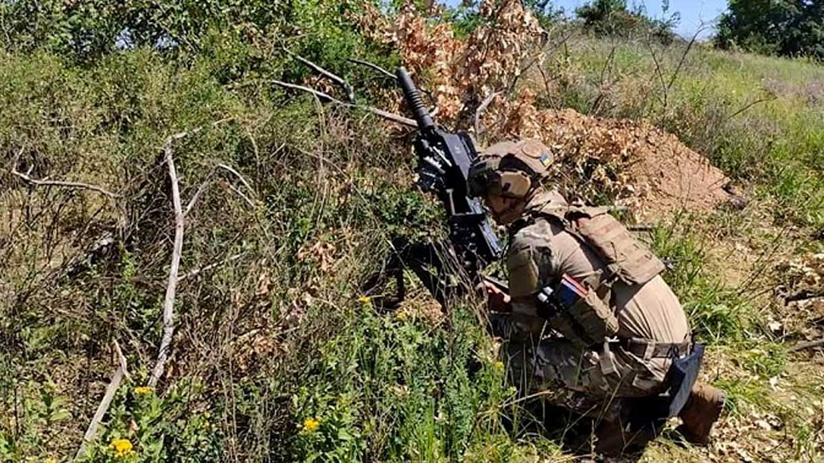 A Ukrainian soldier is operating an automatic grenade launcher while conceal behind some brush and trees. He is well equipped and in a summer uniform during the daytime, crouched down by the weapon