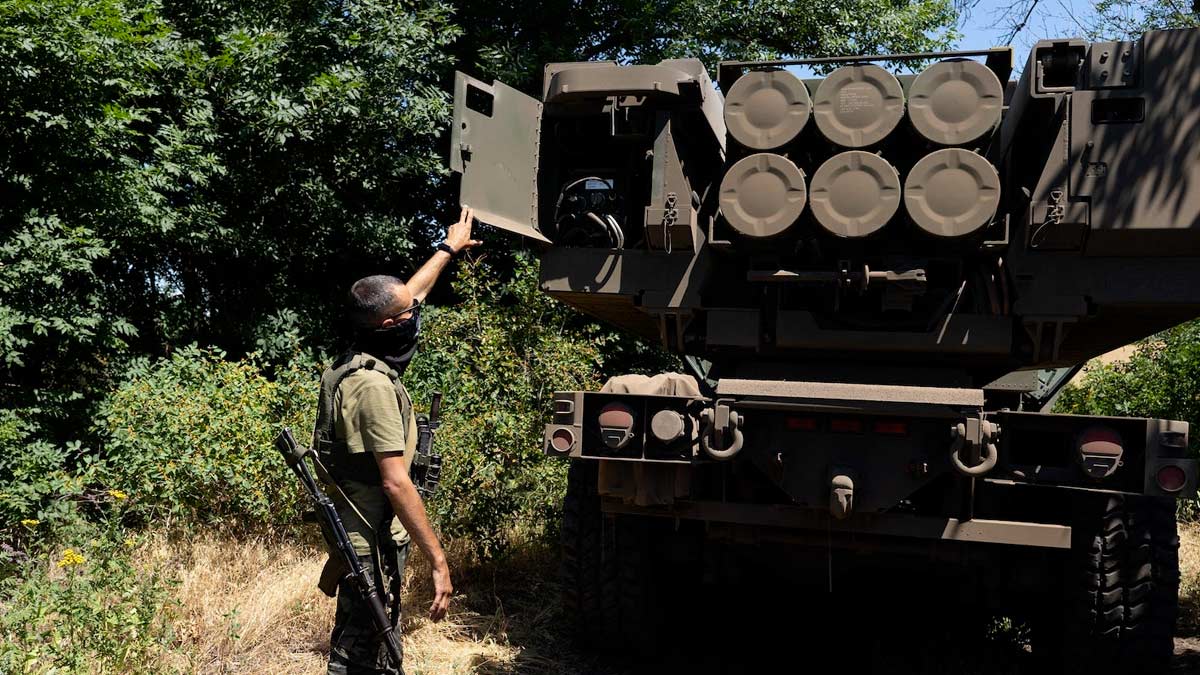 A Ukrainian soldier stands at the rear of a M142 GMLRS HIMARS launcher. The rear doors to the launcher is open and six rockets are loaded. The man is trim, has a rifle slung over his shoulder, and wears a military clothing with his face partially concealed