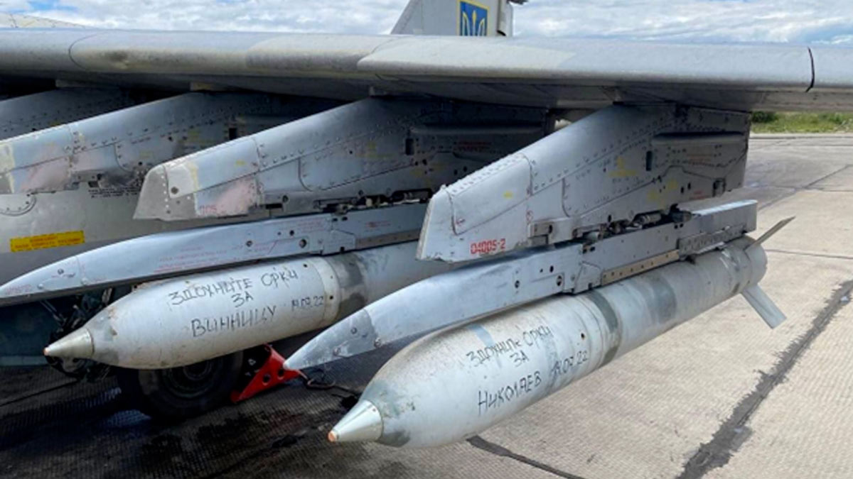 A close up picture shows the wing of a Ukraine Air Force Su-25 ground attack aircraft in gray livery. The aircraft is equipped with S-24 air-to-surface unguided missiles. Written in black marker is "For Mykolaiv" and "For Vinnytsia" on each of the missiles