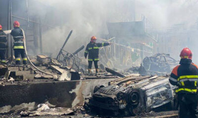 Four firefighters are at a scene of destruction in a Ukrainian city. A car is in the foreground turned on its roof and burned after a missile strike, another car is in the background and through the smoke destroyed since and two story office buildings can be seen. Two fire fighters on the left are fighting a fire out of the picture with water from a hose. A third fire fighter is pointing to something at the right, with a fourth in the foreground, their back turned to the camera. The scene is in grays and browns despite it being early afternoon on a sunny day