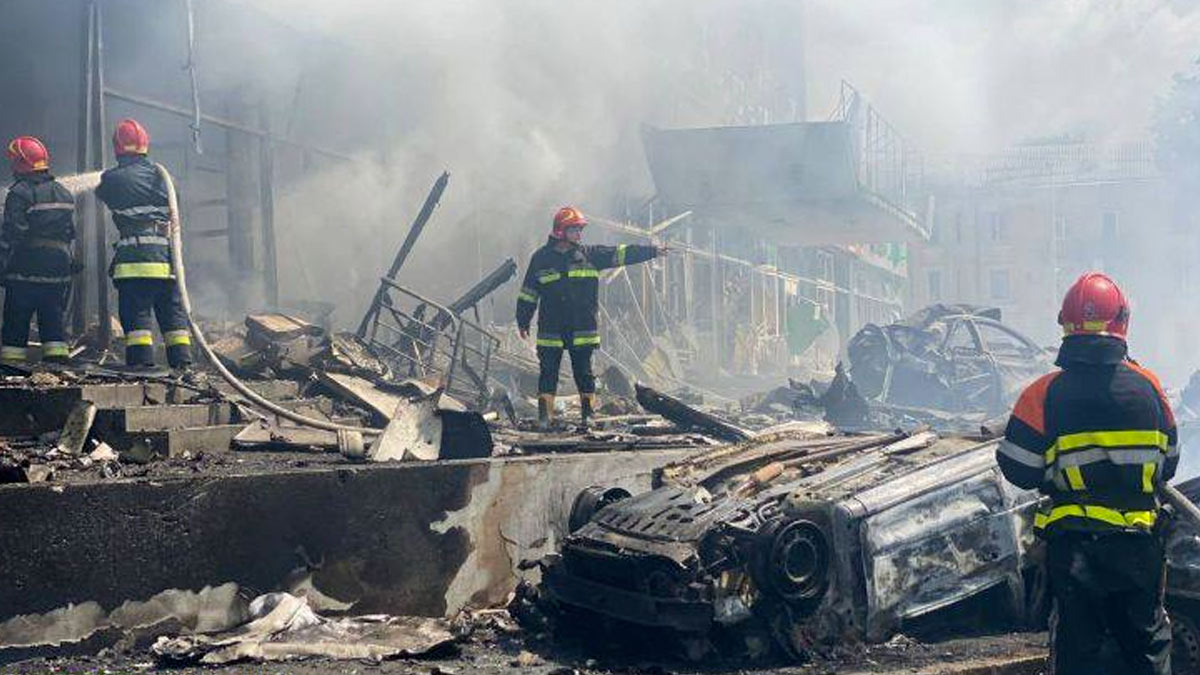 Four firefighters are at a scene of destruction in a Ukrainian city. A car is in the foreground turned on its roof and burned after a missile strike, another car is in the background and through the smoke destroyed since and two story office buildings can be seen. Two fire fighters on the left are fighting a fire out of the picture with water from a hose. A third fire fighter is pointing to something at the right, with a fourth in the foreground, their back turned to the camera. The scene is in grays and browns despite it being early afternoon on a sunny day