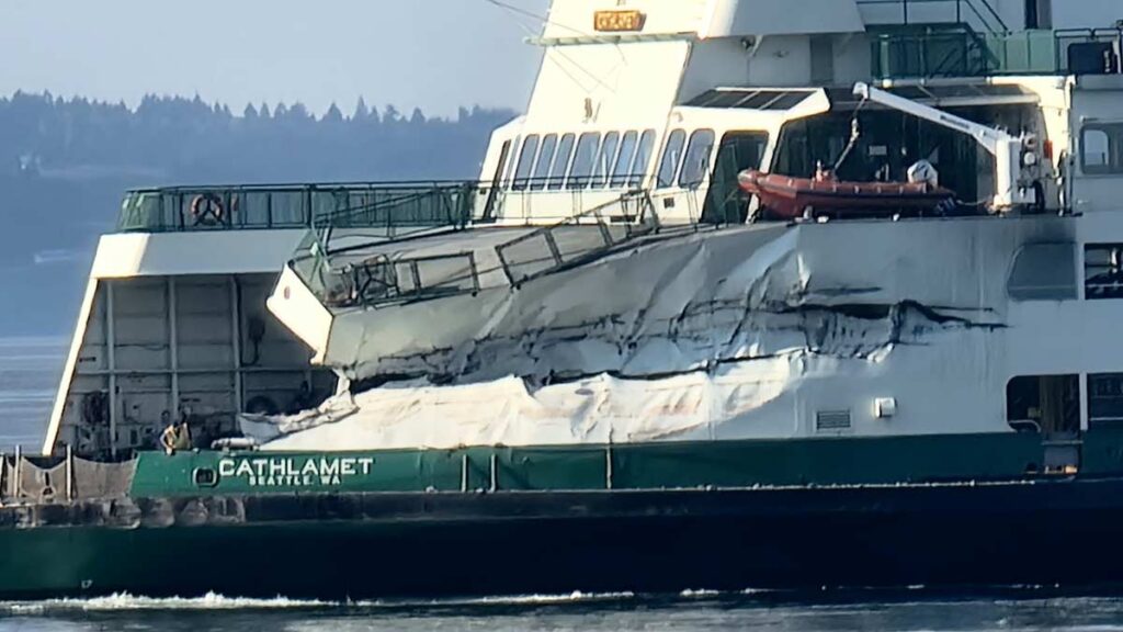 The Washington State Ferry Cathlamet is badly damaged with a large gash in the super structure and the upper part of the ship partially collapsed in the bow area. A ferry worker can be seen peering over the side
