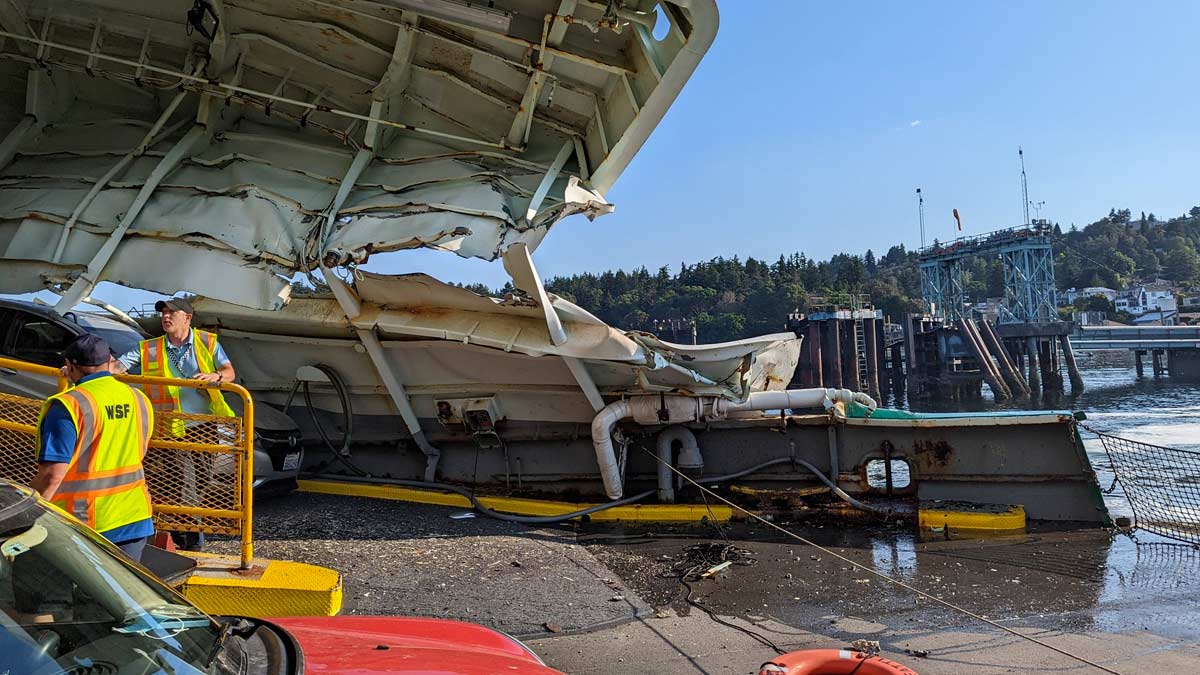 The Washington State Ferry Cathlament is significantly damaged from a hard landing in West Seattle. The picture shows the superstructure between the first and second car deck gashed and crushed, with the second deck where the pickle fork is located collapsed. A silver Honda appears damaged on the ramp between the first and second deck, while crew members manage the emergency