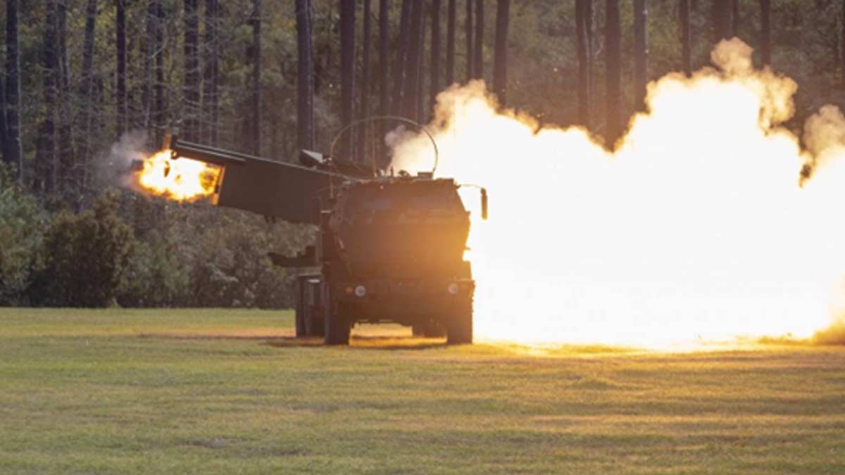 A NATO provided M142 GMLRS launcher, also know as HIMARS is pictured during the day firing. The view is head on, facing the front of the vehicle with smoke and fire coming out from the right side and a rocket firing from the left. The launcher is on a field of short green grass with a field behind it