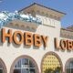 A file photo of the exterior of a Hobby Lobby store with a water bottle superimposed on the left side. Water is coming out of the top of the bottle, framing the left and top of the Hobby Lobby Logo