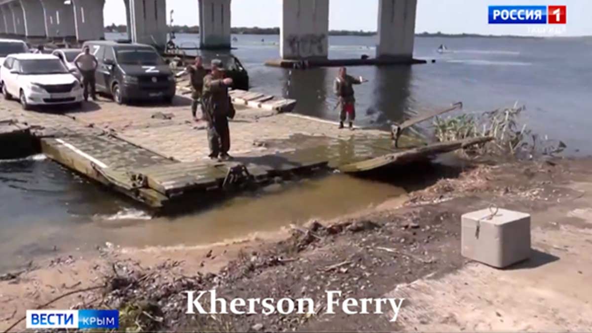 A still image from a news report from Russia 1 in Moscow, shows the military ferry in Kherson mixing military and civilian traffic together. Soldiers operate the ferry, which appears to be arriving and preparing to unload. A Russian military van marked with Z is at the wrong, while a civilian sedan is waiting to unload to its left