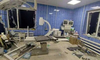 A trauma and exam room with blue tile walls and medical equipment is in ruins after a missile attack on the Mykolaiv hospital - the windows are broken with one window frame blown into the room, the blast force damaged equipment and push it to one side. The electricity still works and the lights are on, it is night outside