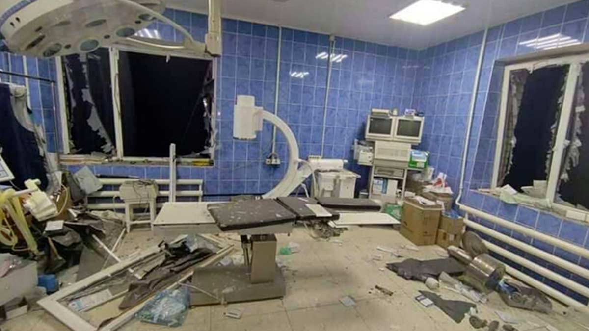 A trauma and exam room with blue tile walls and medical equipment is in ruins after a missile attack on the Mykolaiv hospital - the windows are broken with one window frame blown into the room, the blast force damaged equipment and push it to one side. The electricity still works and the lights are on, it is night outside