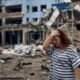 A woman with red hair wearing a striped shirt that is black and white, holds her had and looks down in sadness - behind her is a bombed out building in the city of Mykolaiv that has suffered heavy damage