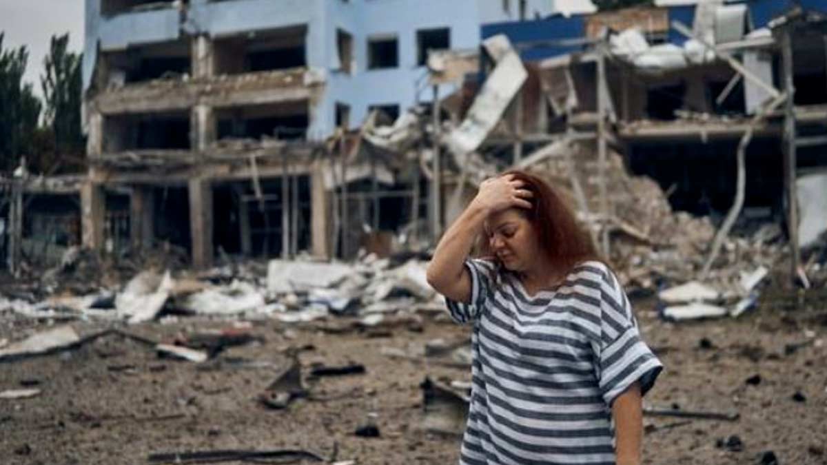 A woman with red hair wearing a striped shirt that is black and white, holds her had and looks down in sadness - behind her is a bombed out building in the city of Mykolaiv that has suffered heavy damage