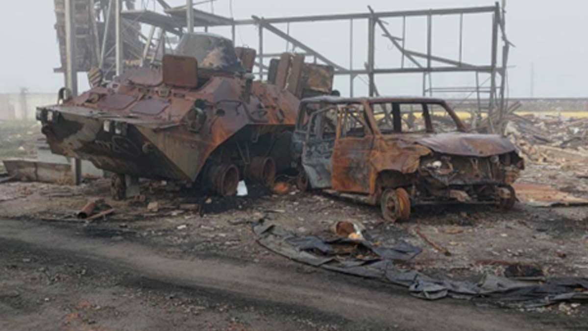 The burned hulk of a Russian armored command vehicle and a SUV are in the rubble of a destroyed warehouse in Nova Kakhova - both vehicles are completely destroyed and only the metal frame of a building remains - smoke still hangs in the air from the rocket attack giving the picture sepia tones