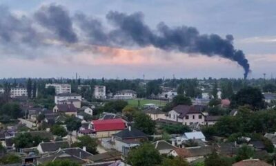 Black smoke rises from the horizon with the wind carrying it from right to left in morning light and a cloudy sky. The homes and buildings of Russia-occupied Crimea are in the foreground