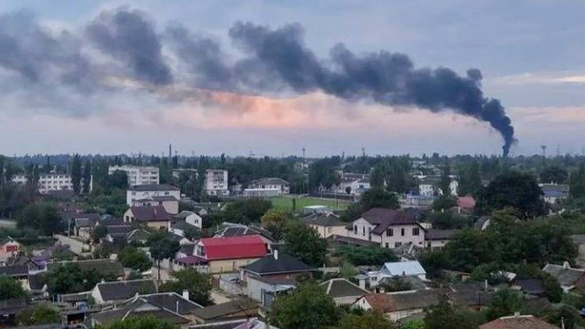Black smoke rises from the horizon with the wind carrying it from right to left in morning light and a cloudy sky. The homes and buildings of Russia-occupied Crimea are in the foreground