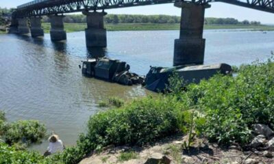 Two Russian supply trucks are attempting to drive across the Oskil River next to a disabled bridge near Borova - the trucks and laden and one is pulling a trailler. The day is sunny, and the bridge is damaged on both sides. The water is up to the cab on the lead truck, which is still close to the riverbank. In the foreground is a person in a white shirt sitting on the river bank watching the trucks.