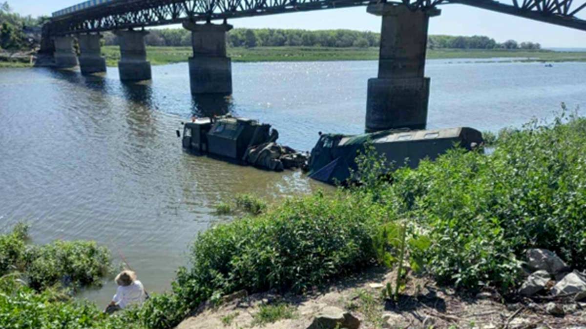 Two Russian supply trucks are attempting to drive across the Oskil River next to a disabled bridge near Borova - the trucks and laden and one is pulling a trailler. The day is sunny, and the bridge is damaged on both sides. The water is up to the cab on the lead truck, which is still close to the riverbank. In the foreground is a person in a white shirt sitting on the river bank watching the trucks.