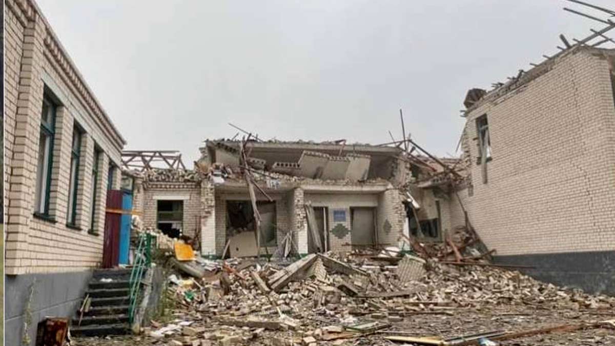 The inside of a courtyard of a light two story brick building destroyed by artillery. The second floor has collapsed and the outside wallk is destroyed, showing the interior rooms. The courtyard is full of debris and the sky is cloudy