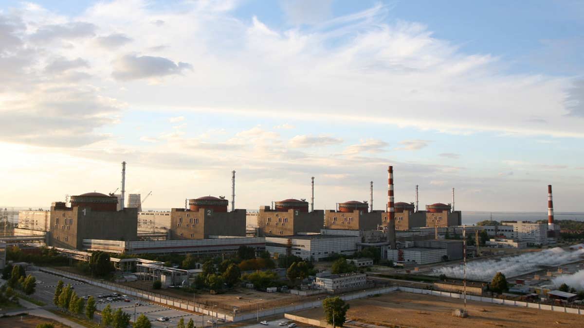A photo of the Zaporizhzhia Nuclear Power Plant before the war in morning light, looking at the six reactor buildings. The Dnipro River can be seen in the background, the sky has high clouds